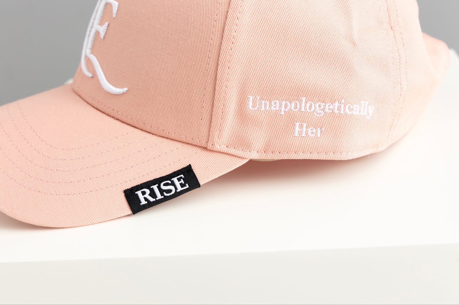 Pink cap with 'RISE' and 'Unapologetically Her' text on a light background